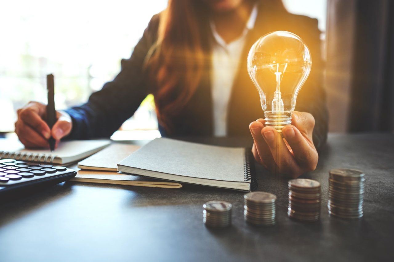 Businesswoman holding a lightbulb while taking note on notebook with coins stack on table, saving energy and money concept